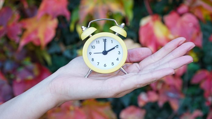 A woman holds an alarm clock against the background of autumn leaves. Reminder to switch to winter time.