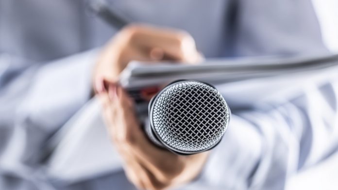 A journalist holds a microphone at a press conference and writes information in a notebook.
